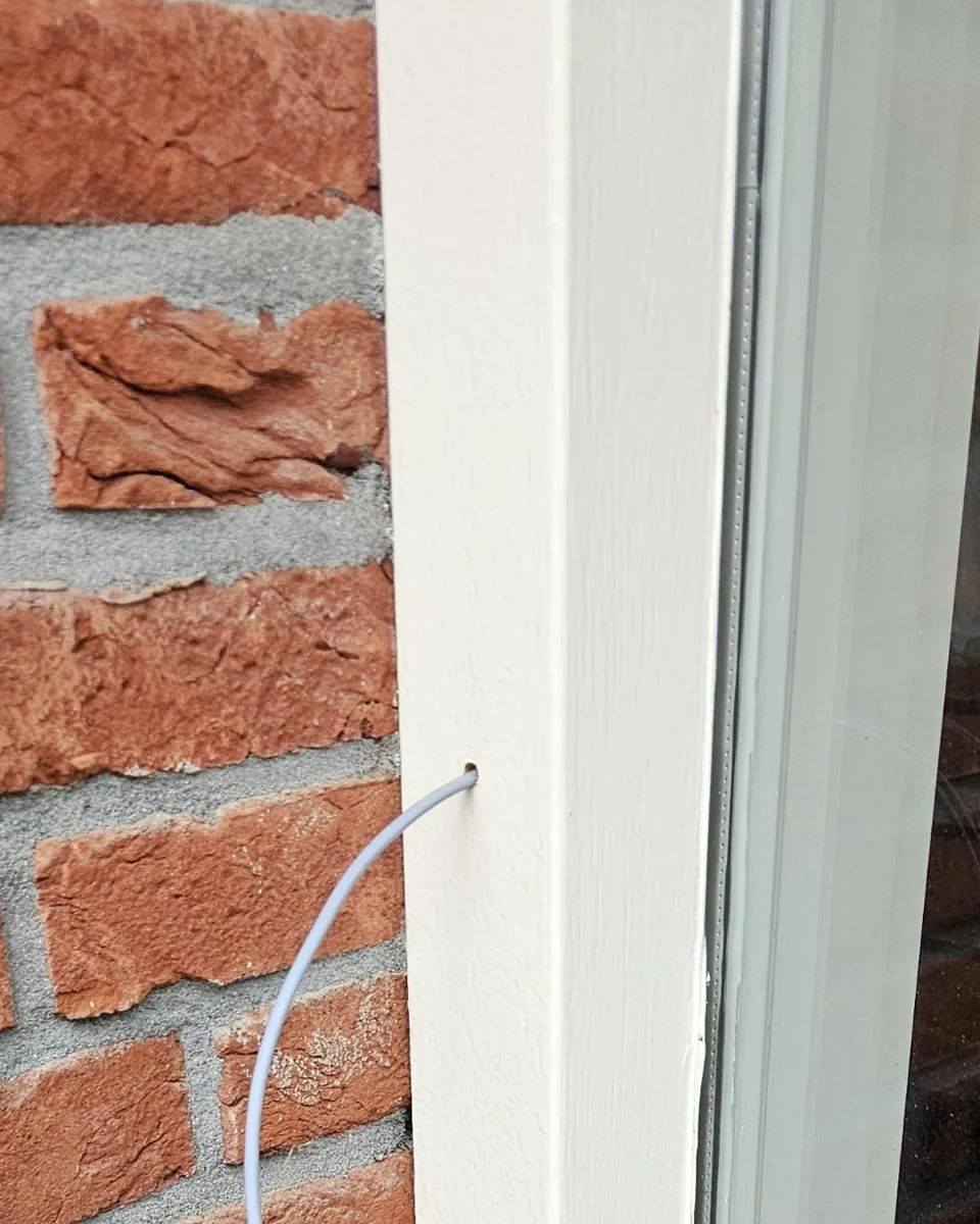 Een dunne witte kabel komt uit een gaatje in het witte kozijn naast rode baksteen