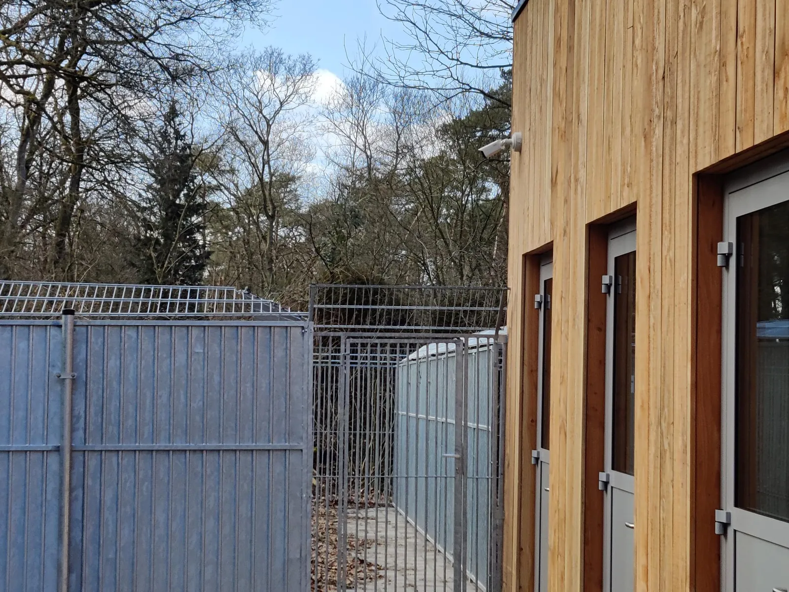Witte bulletcamera aan een houten gevel, gericht op de buitenrennen en het hekwerk van een dierenopvang