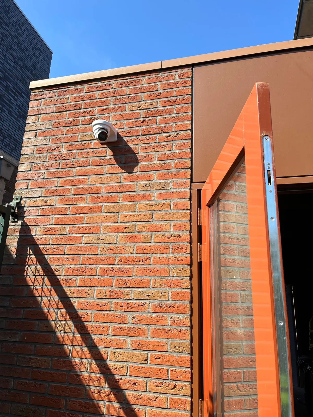 Camera hoog op een oranjerode zijgevel naast een open poortdeur in Almere, met harde schaduwen