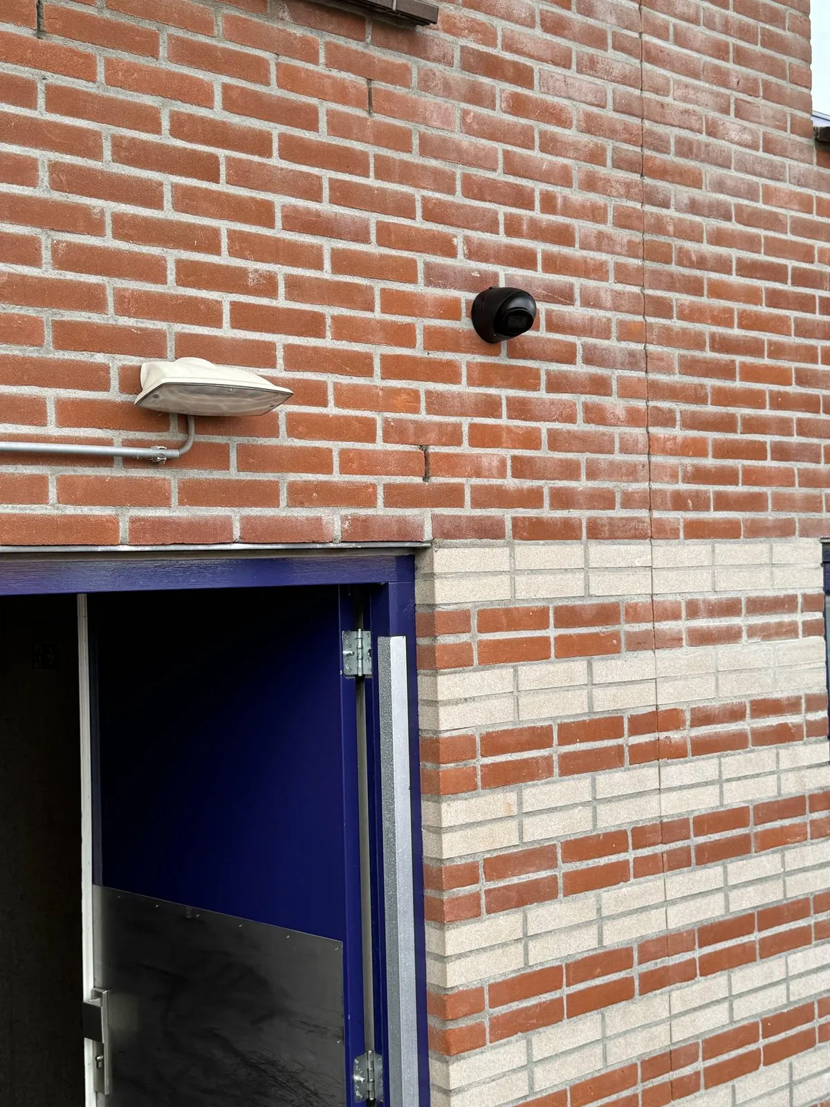 Camera boven de blauw geschilderde nooddeur in Ede, naast de buitenlamp op het metselwerk