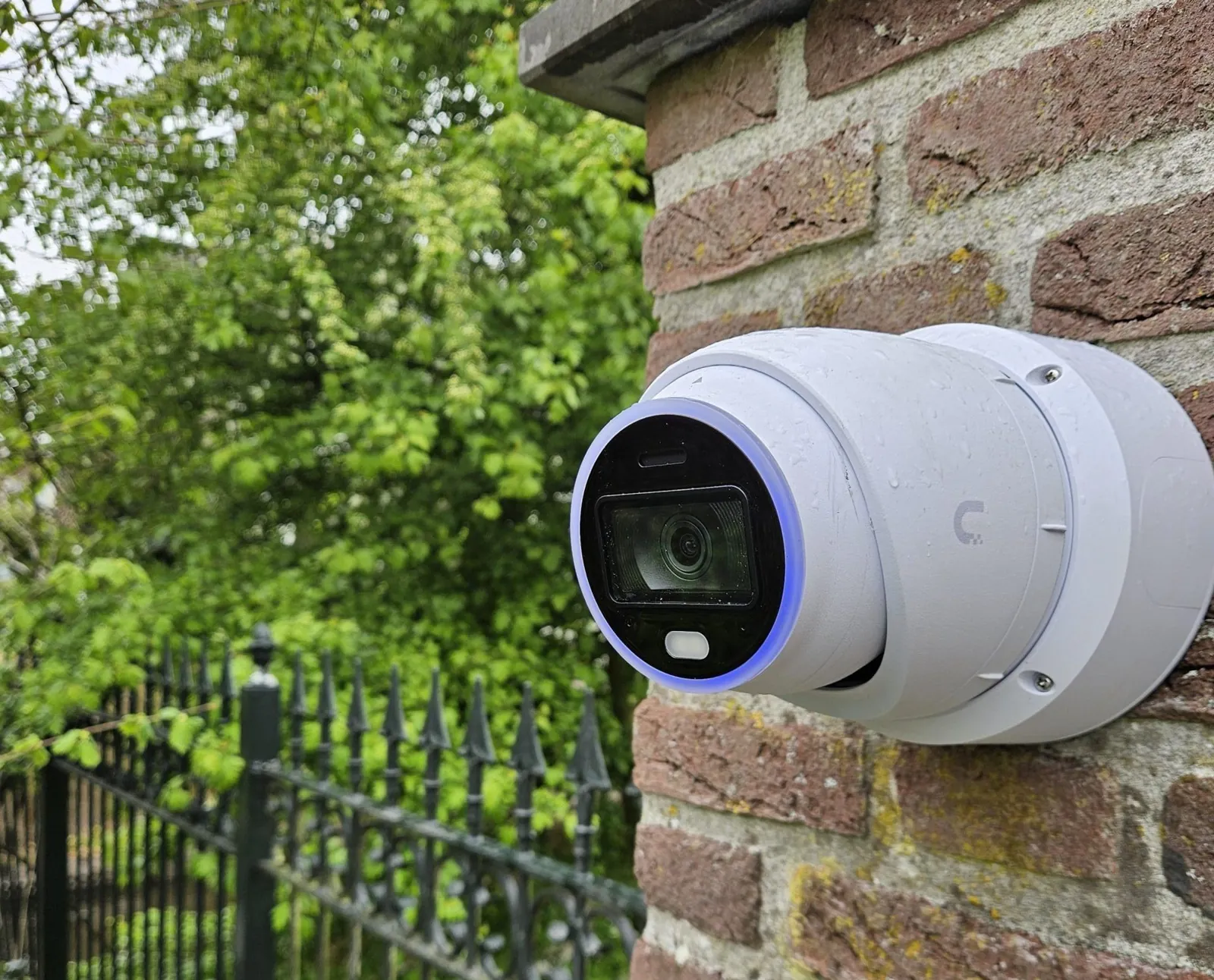 Witte camera op de bakstenen pilaar van een toegangspoort bij een woning, met sierhekwerk en groen erachter