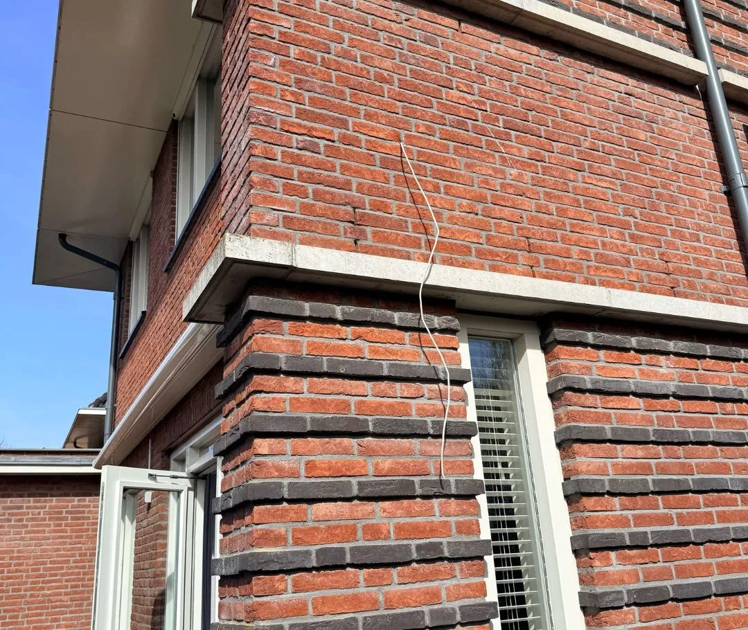 Camerakabel strak omlaag langs de hoek van het metselwerk, richting het raam
