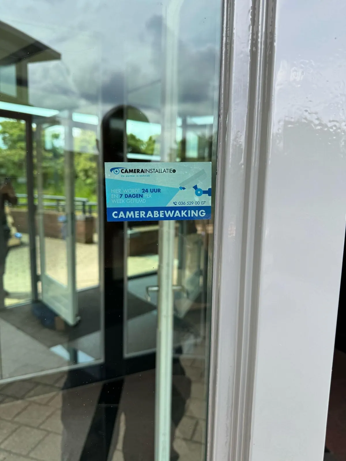 Camerasticker op de glazen toegangsdeur in Ede, met de overkapping weerspiegeld