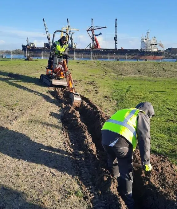 Minigraver trekt een kabelsleuf door het gras van een haventerrein in Amsterdam, met kranen en een schip op de achtergrond