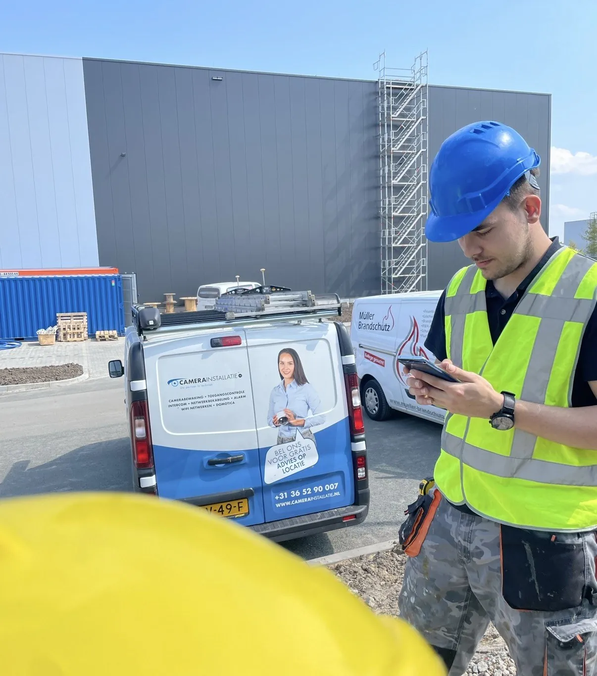 Monteur in helm en hesje leest de bouwtekening op zijn tablet, met onze bus erachter