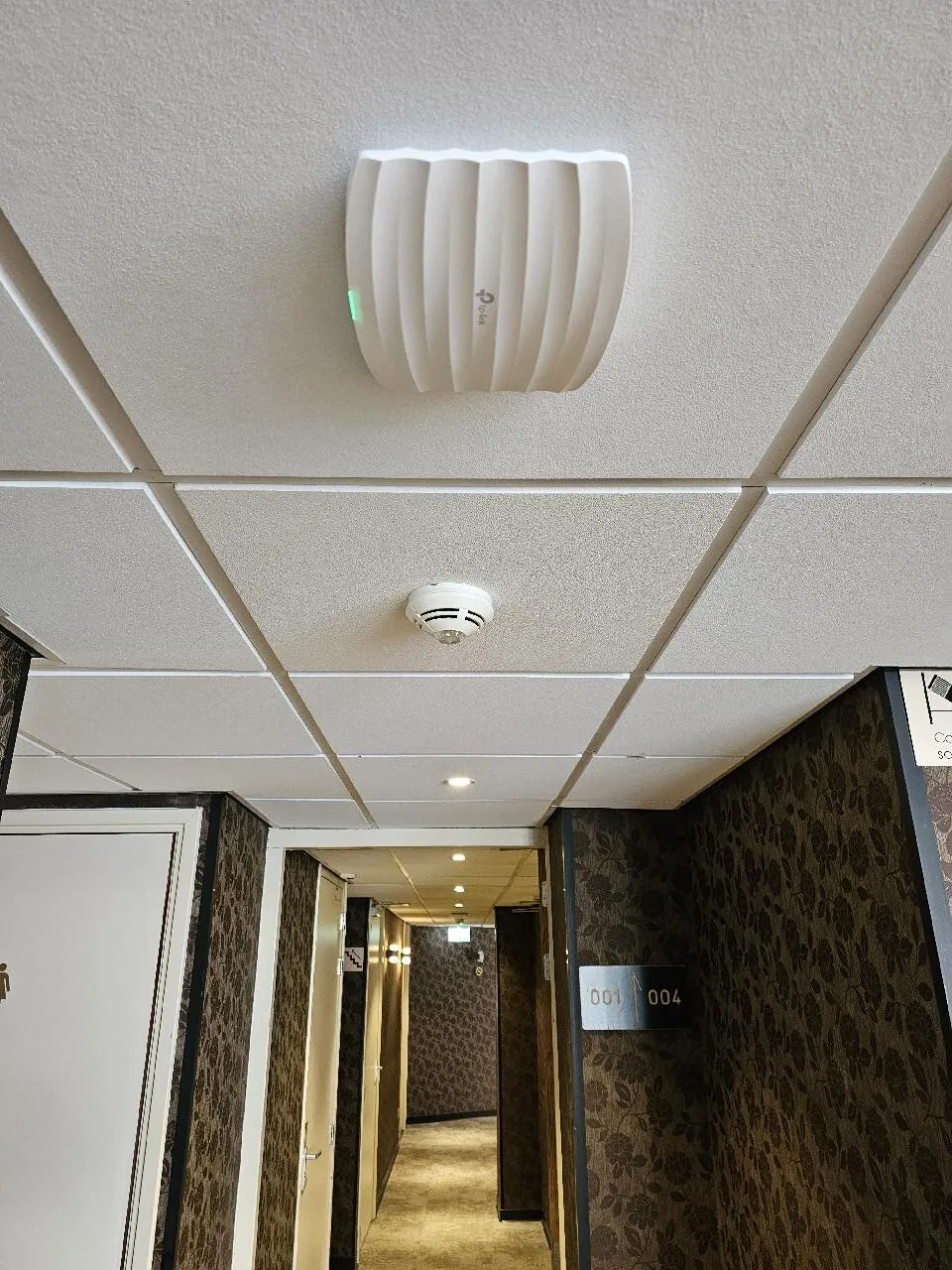 Wit Omada-access point aan het systeemplafond van een hotelgang in Amsterdam, met kamerdeuren erachter