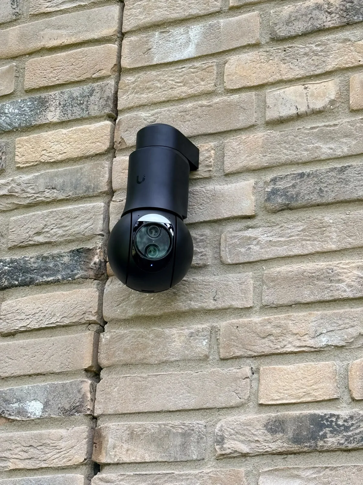Close-up van de zwarte UniFi PTZ op een muurbeugel tegen baksteen in Voorschoten