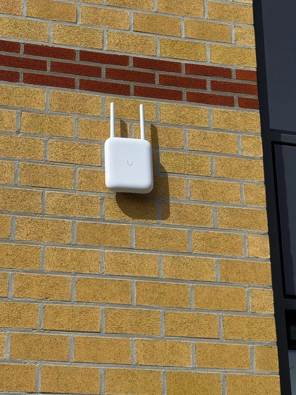 Wifi-zender van UniFi met twee antennes buiten op de geel gemetselde gevel van een bedrijf