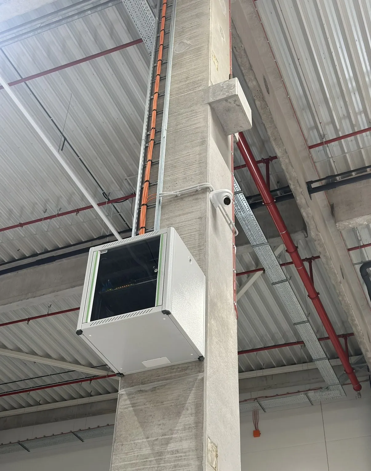 Wandpatchkast en domecamera aan een betonnen kolom in een vrachthal, met oranje glasvezel in de goot